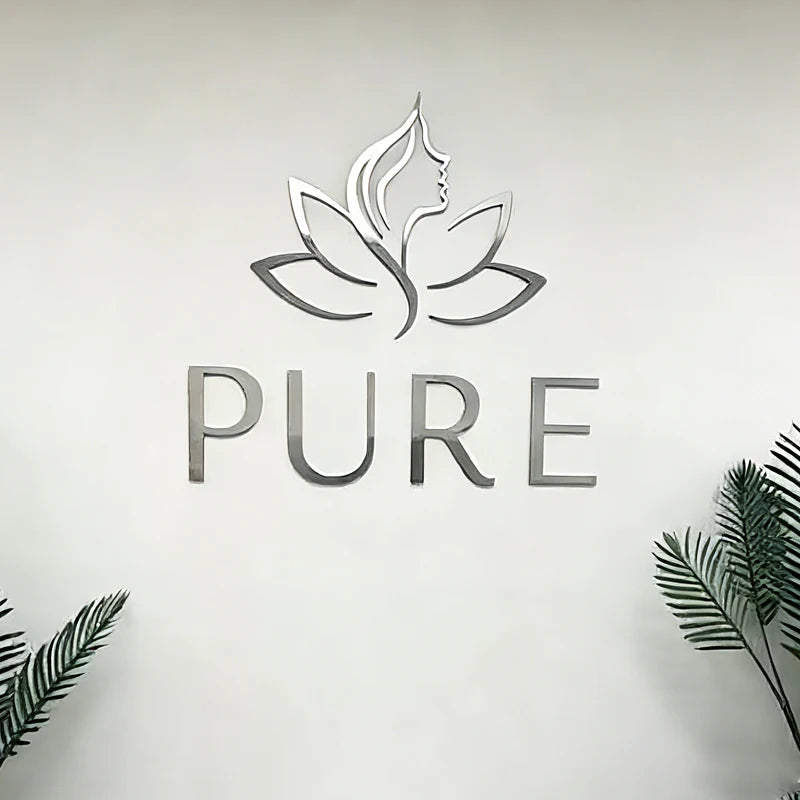 Silver metal wall art featuring a lotus flower with a woman's profile and the word 'PURE' below it, mounted on a white wall with decorative green leaves at the bottom corners.