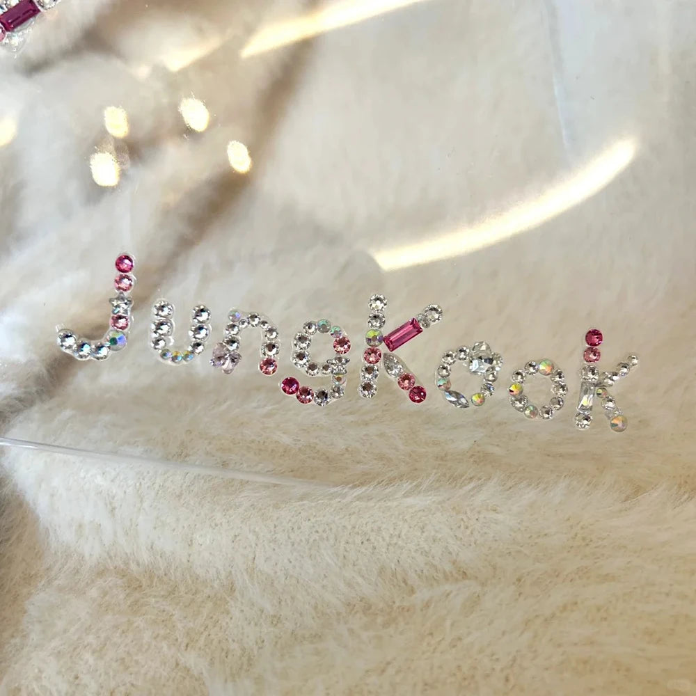 A rhinestone nameplate spelling 'Jungkook' in silver letters, accented with pink and iridescent crystals, resting on a soft beige faux fur surface.
