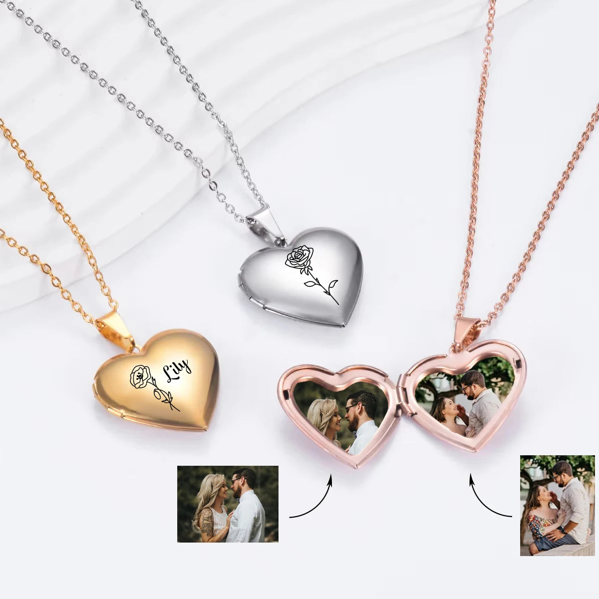 Three heart-shaped pendant necklaces are displayed on a white draped fabric background: one gold, one silver, and one rose gold. The gold and silver pendants are closed and engraved with names and a rose motif.