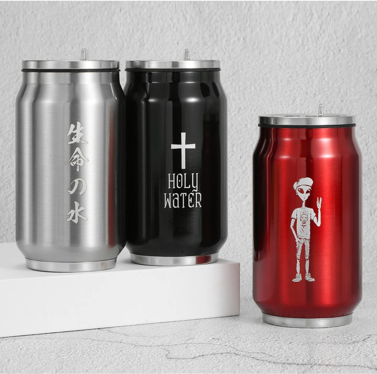 Three stainless steel can-shaped cups with lids are displayed on a white riser and a light gray surface. The left cup is silver with vertical Japanese characters, the middle cup is black with a white cross and the words 'HOLY WATER,' and the right cup is red with a white alien graphic.