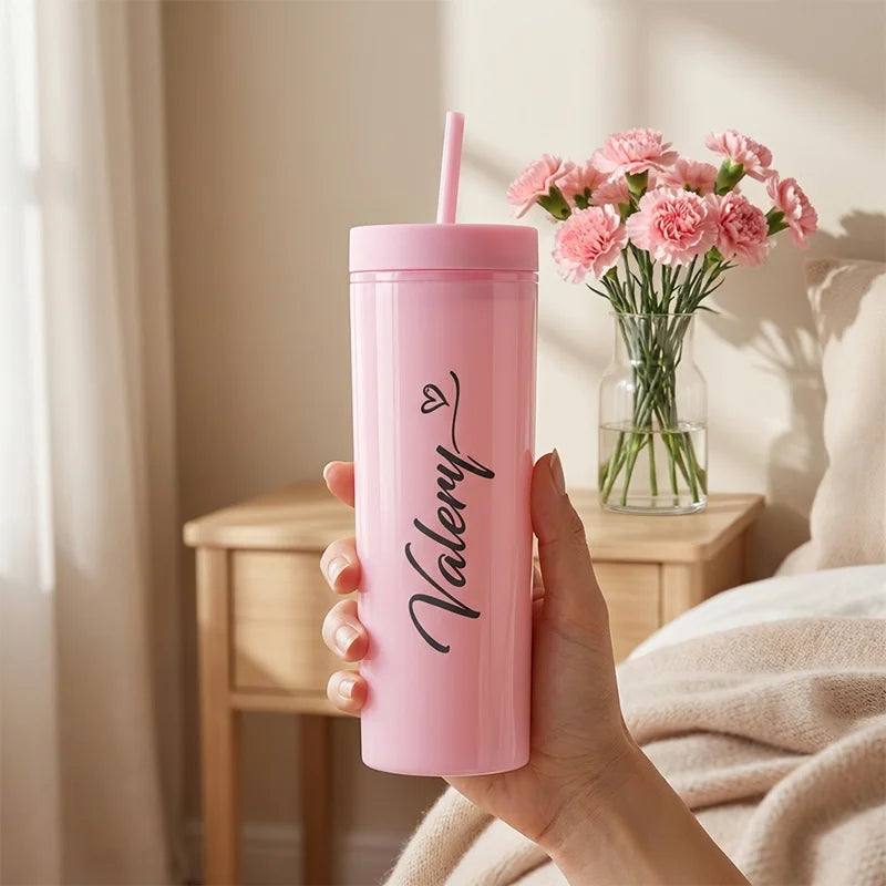 Pink tumbler with straw held by a hand, next to a vase of pink flowers on a wooden table.