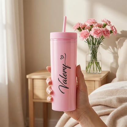 Pink tumbler with straw held by a hand, next to a vase of pink flowers on a wooden table.