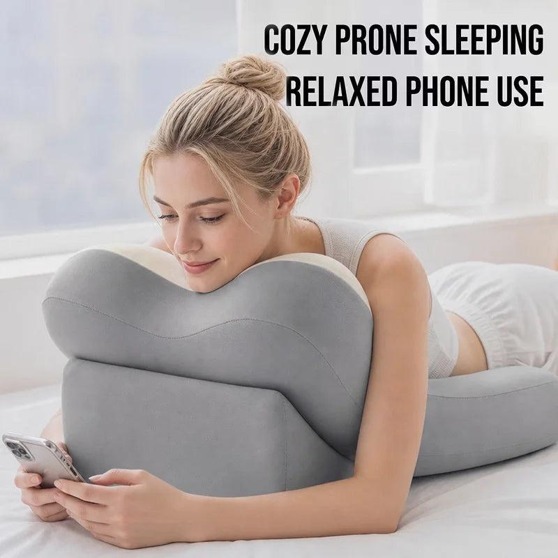A woman lying on a bed uses a gray U-shaped phone pillow while holding a smartphone. Text at the top reads 'COZY PRONE SLEEPING RELAXED PHONE USE'.