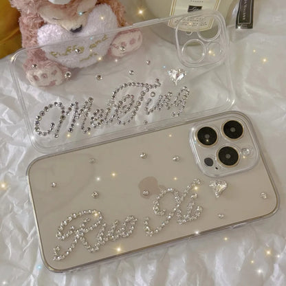 Two clear phone cases decorated with rhinestones and cursive text, displayed on a white textured surface with a plush toy in the background.