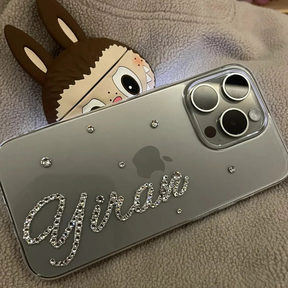 A silver iPhone case decorated with rhinestone script reading 'Ayan' and small rhinestone dots, shown resting on a beige textured surface with a cartoon llama prop peeking out from behind.