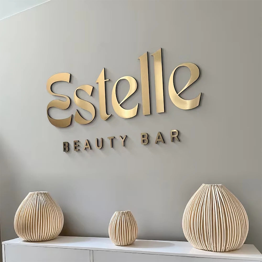 Gold 'Estelle Beauty Bar' sign on a gray wall with decorative vases below.