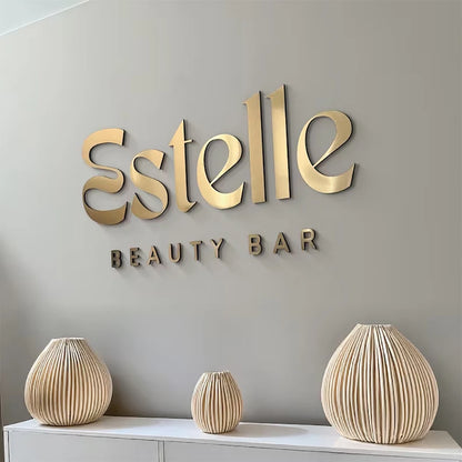 Gold 'Estelle Beauty Bar' sign on a gray wall with decorative vases below.