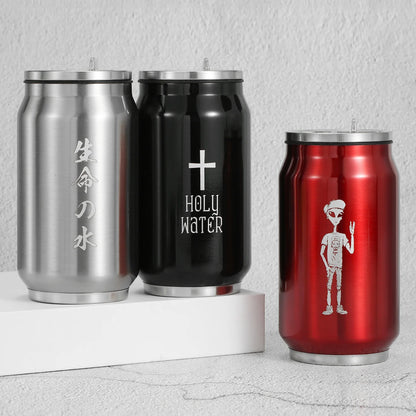 Three stainless steel can-shaped cups with lids are displayed on a white riser and a light gray surface. The left cup is silver with vertical Japanese characters, the middle cup is black with a white cross and the words 'HOLY WATER,' and the right cup is red with a white alien graphic.