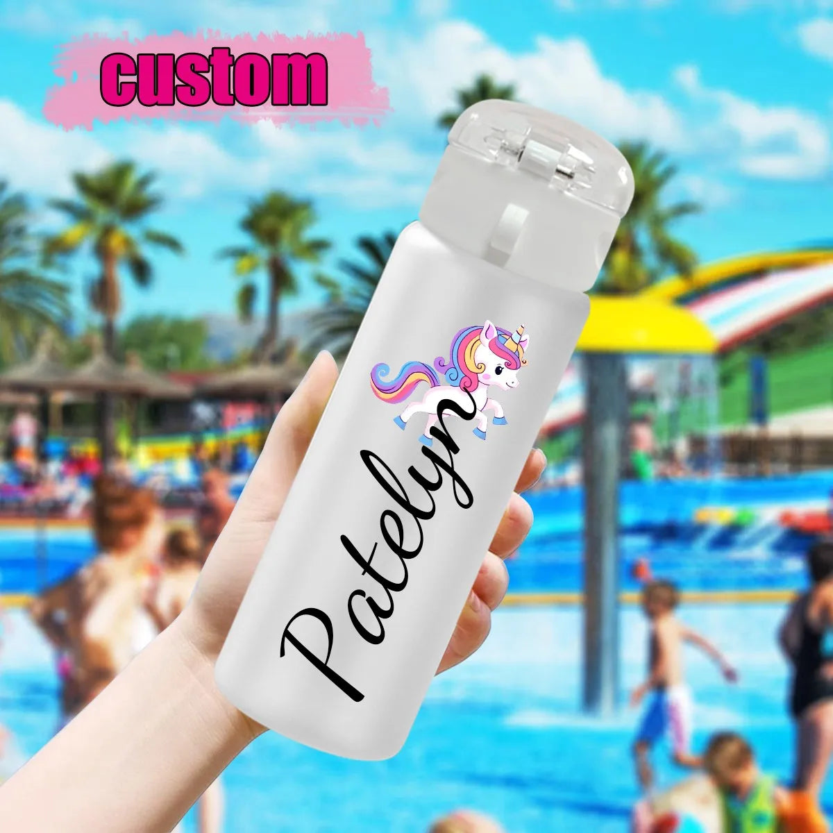 A hand holds a white insulated water bottle with a flip-top lid at a poolside location. The bottle is printed with a colorful unicorn illustration and the name 'Patelyn' in black script.