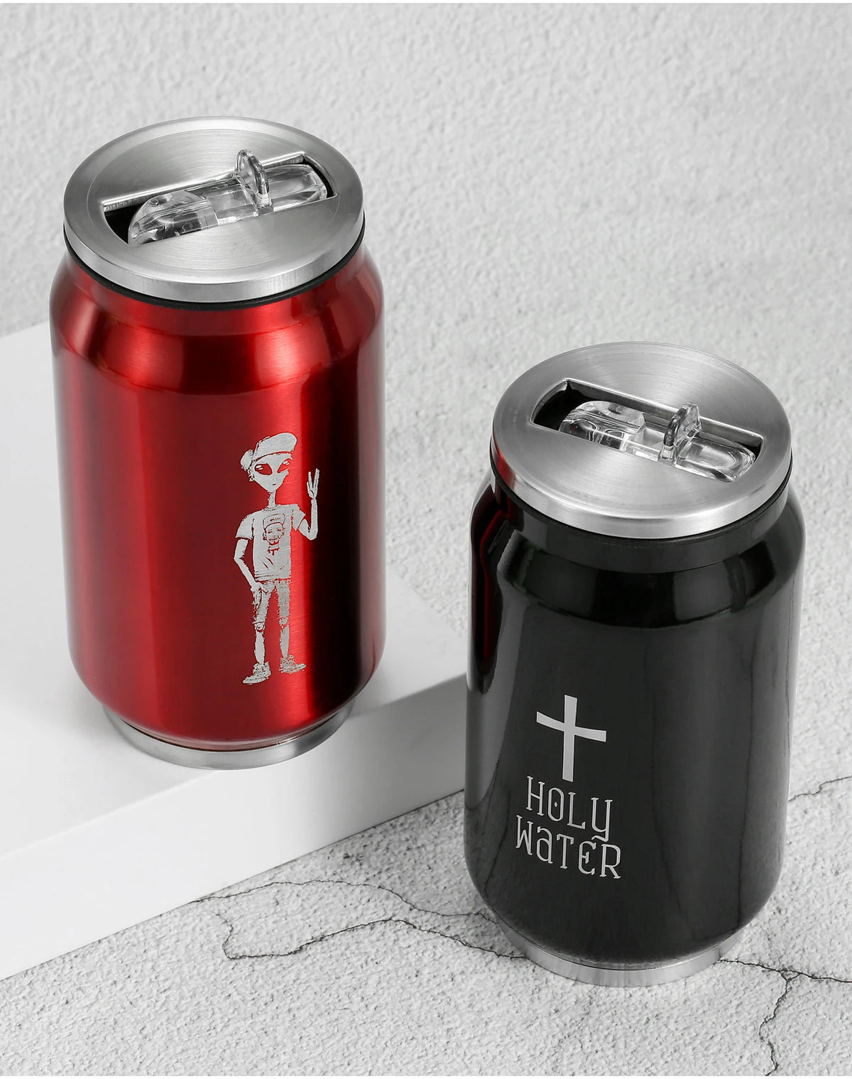 Two slim aluminum tumbler cans with flip-top lids, shown on a light textured surface. The left can is metallic red with a silver alien graphic, and the right can is black with a silver cross and the words 'HOLY WATER'.