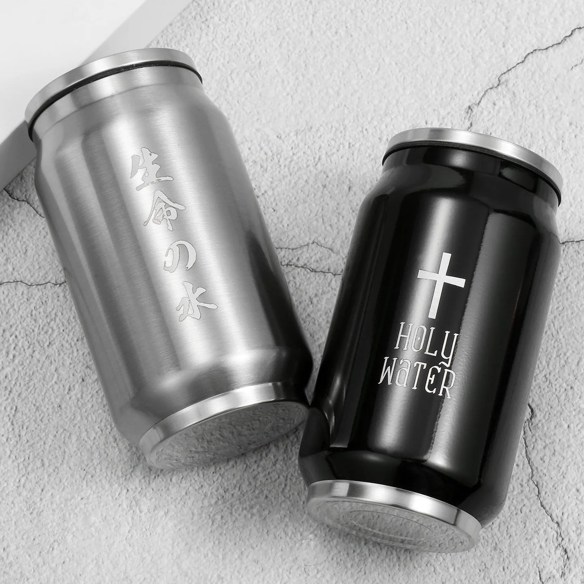 Two stainless steel can-shaped tumblers on a light textured surface. One tumbler is brushed silver with vertical Japanese characters, and the other is black with a white cross and the words 'HOLY WATER'.