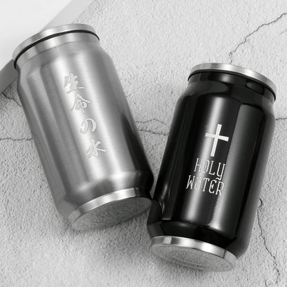 Two stainless steel can-shaped tumblers on a light textured surface. One tumbler is brushed silver with vertical Japanese characters, and the other is black with a white cross and the words 'HOLY WATER'.