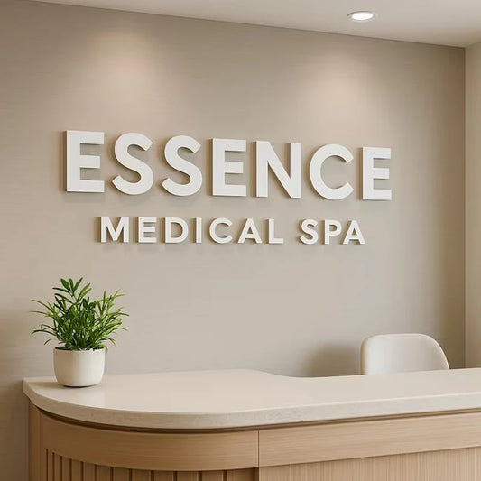 A modern reception desk in a spa setting with large white wall lettering reading 'ESSENCE MEDICAL SPA' above it. A small potted green plant sits on the white countertop, and a white chair is visible against a warm beige wall.